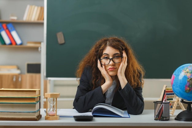 triest greep het hoofd van een jonge vrouwelijke leraar met een bril die aan het bureau zit met schoolhulpmiddelen in de klas