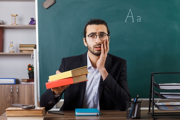 Triest hand op de wang leggen mannelijke leraar met een bril die een boek vasthoudt bij de camera die aan tafel zit met schoolhulpmiddelen in de klas