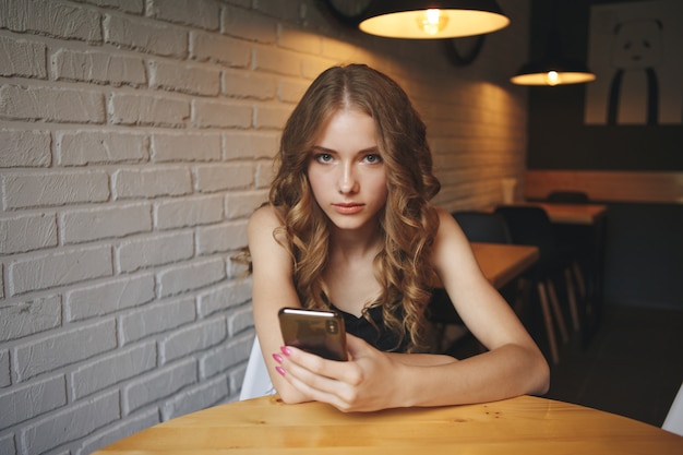 Triest jong meisje zit in een café op zoek moe haar telefoon, ongelukkig meisje kijkt naar haar zwarte telefoon,