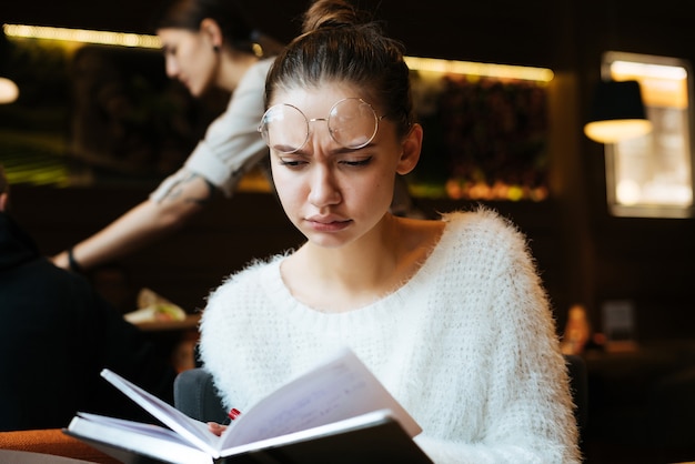 Triest jonge studente in glazen en in witte trui geeft lezingen in café