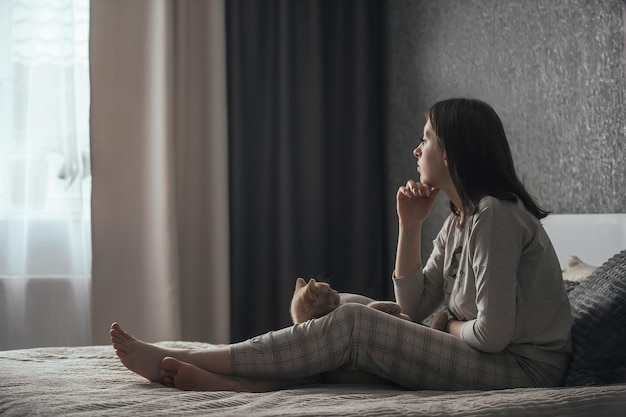 Triest jonge vrouw in pyjama met seizoensgebonden affectieve stoornis zittend op bed voor raam