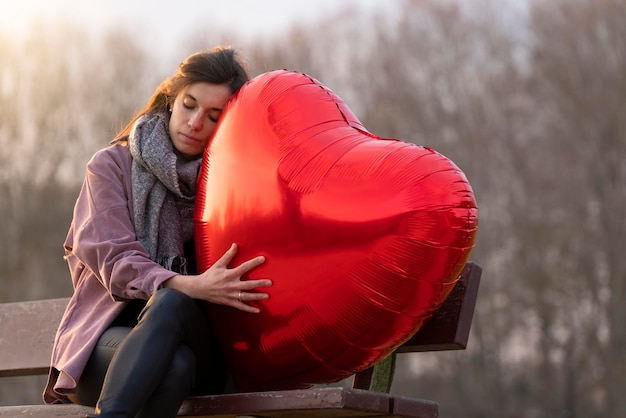 Triest jonge vrouw knuffelen een hartvormige ballon zittend op een bankje in het park en denken met zijn ogen dicht