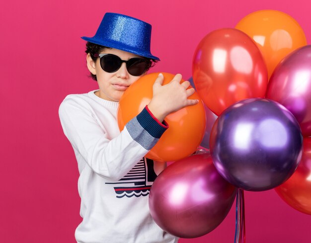 Foto triest jongetje met een blauwe feestmuts met een bril die ballonnen vasthoudt die op een roze muur zijn geïsoleerd