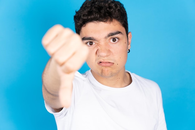 Triest negatieve Spaanse tiener jongen duimen naar beneden kijken naar camera geïsoleerd op blauwe achtergrond