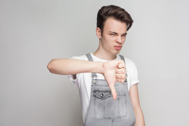 Triest ontevreden jonge brunette man in casual stijl met wit t-shirt en denim overall staan en kijken naar camera met duimen naar beneden en verdriet. indoor studio opname, geïsoleerd op een grijze achtergrond.