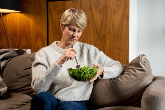 Triest vrouw van middelbare leeftijd eet een smakeloze groene groentesalade zittend op de bank