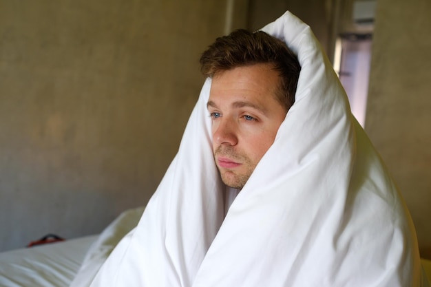 Trieste blanke man zittend op zijn bed met zijn hoofd bedekt met deken