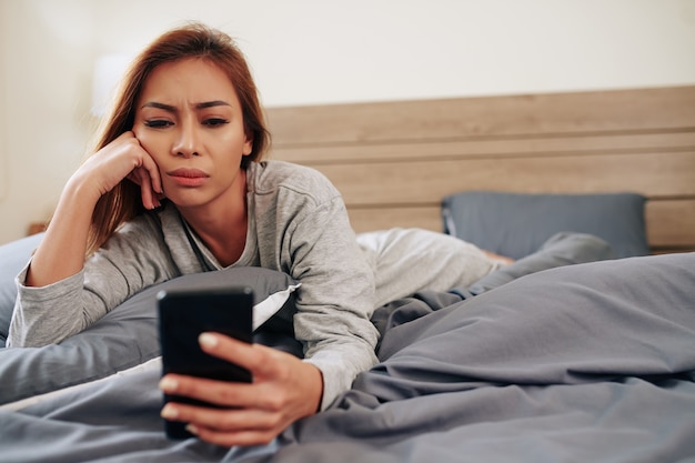 Trieste emotionele jonge vrouw die slecht nieuws leest op het scherm van de smartphone als ze in bed ligt