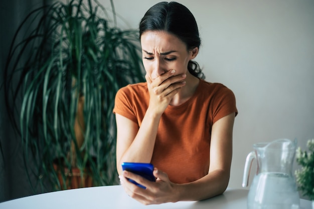 Trieste, gefrustreerde jonge brunette vrouw huilt met smartphone in handen terwijl ze op de stoel in het appartement zit