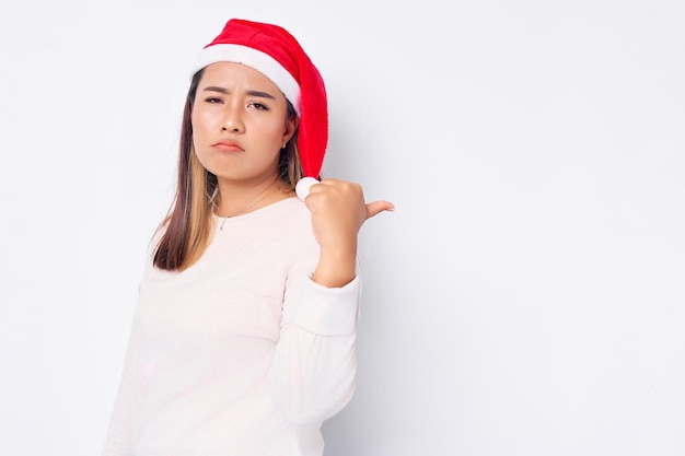 Trieste jonge Aziatische vrouw in een kerstman hoed wijzende duim op kopieerruimte geïsoleerd op witte achtergrond viering Kerst vakantie en Nieuwjaar concept