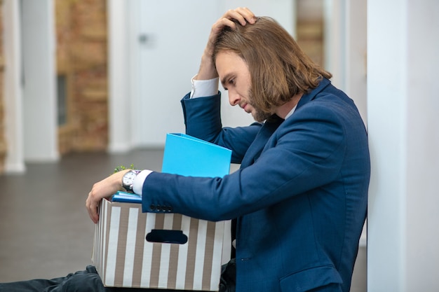 Foto trieste jonge volwassen man in pak zittend met doos op vloer binnenshuis hand op hoofd te houden