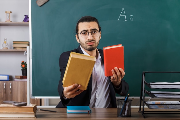 Trieste mannelijke leraar met een bril die een boek vasthoudt dat aan tafel zit met schoolhulpmiddelen in de klas