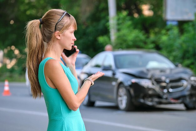 Trieste vrouwelijke chauffeur die spreekt op sellphone aan straatkant en belt voor hulpdiensten na auto-ongeluk. Verkeersveiligheid en verzekeringsconcept.