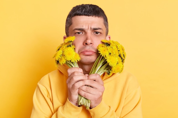 Trieste zieke man met casual stijl hoodie poseren geïsoleerd over gele achtergrond met boeketten van paardebloemen kijkend naar camera met pruillip lippen boos van allergie