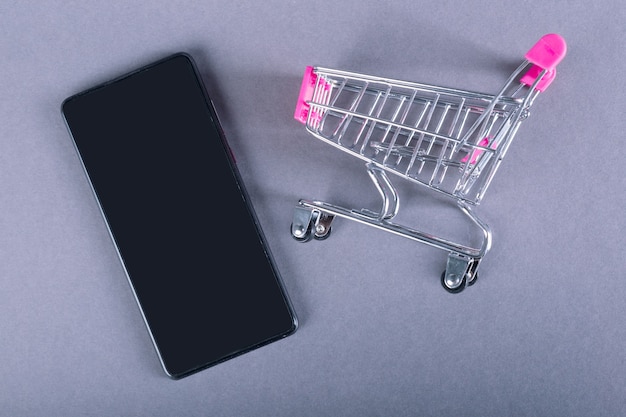 Trolley and black smartphone isolated on gray background. Shopping online concept.