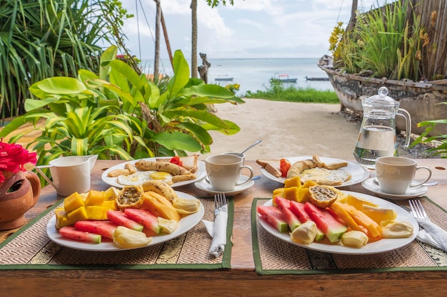 바다 근처 해변에서 2인용 과일 커피와 스크램블 에그, 바나나 팬케이크로 구성된 열대 조식