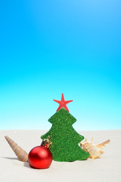 Tropical summer beach Christmas with a Xmas tree, bauble and shells on golden sand and copy space for your greeting on blue sky