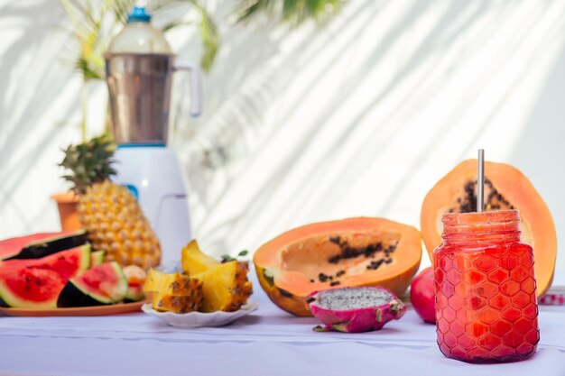 Tropisch fruit Ingrediënten granaatappel, ananas, papaya, dragon fruit, sinaasappel en watermeloen met mixer op witte tafel zomer achtergrond palmtak. twee glazen smoothiepot met strobuis copyspace