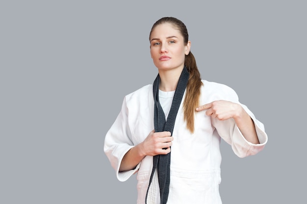 Trotse aantrekkelijke atletische vrouw in witte kimono staande en wikkelt zwarte riem om de nek en wijzende vinger. Japans karate- en sportconcept. indoor studio-opname, geïsoleerd, grijze achtergrond
