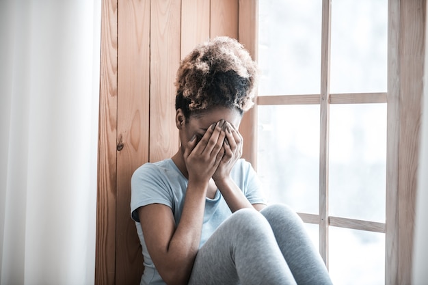 Trouble, sadness. Upset dark skinned woman with curly hair in tshirt covering face with hands sitting near window at home