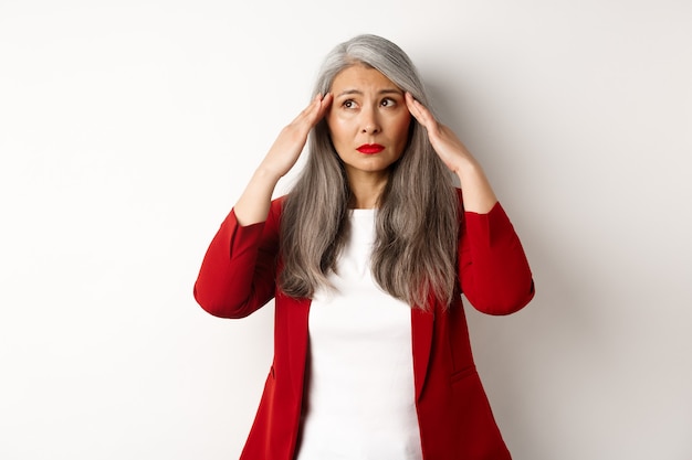 Troubled asian office woman touching head, looking aside and looking alarmed, trying calm down, standing over white background.