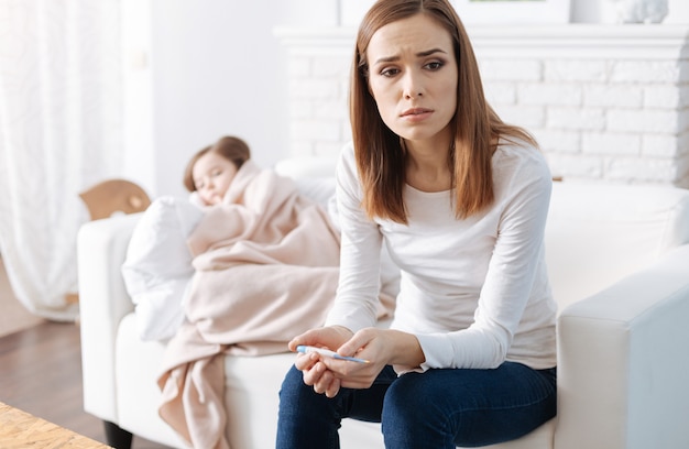 Troubled caring mother holdign thermometer and sitting on the couch while her little sick daughter sleeping in the background