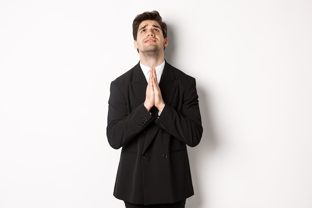 Troubled and hopeful man in black suit begging god, pleading and looking up, need help, standing over white background.