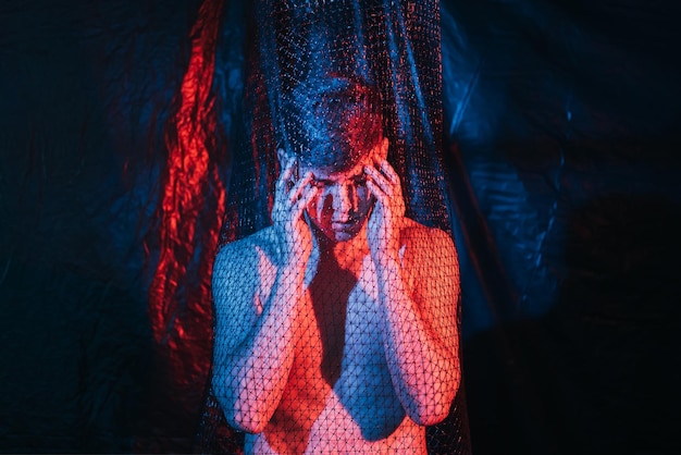 Troubled man. Panic attack. Emotional crisis. Social pressure. Art portrait of paranoid hurt shirtless guy suffering from headache in mesh trap in red blue neon light at night on dark background.