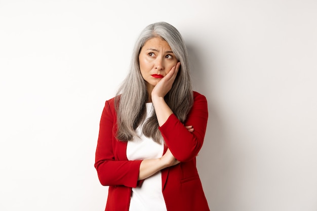 Photo troubled and sad asian female entrepreneur looking at upper left corner, thinking with upset face, standing over white background