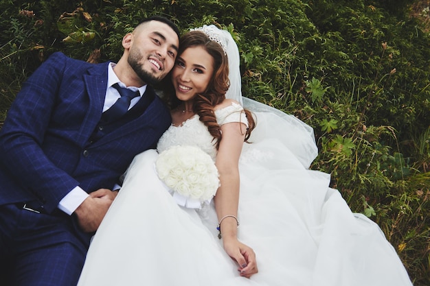 Trouwfoto van een liefdevol Kazachs Aziatisch koppel in de natuur, een mooie bruid in een witte jurk en sluier, en een bruidegom in een pak knuffel en kus