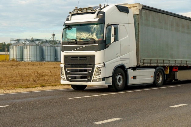 A truck is passing on the road in the background silver silos on agro manufacturing plant for processing drying cleaning and storage of agricultural products flour Large iron barrels of grain