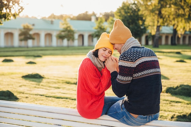 True feelings and romantisism concept Adorable young woman in knitted yellow hat and red warm sweater warms her hands in boyfriends hands sit together on bench closes eyes with great enjoyment