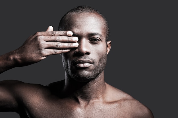 True masculinity. Portrait of young shirtless African man covering one eye with hand and looking at camera while standing against grey background