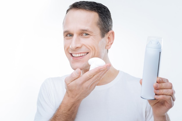 For true men. Positive nice delighted man smiling and holding a shaving foam while preparing for shaving