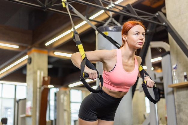 Trx training giovane donna rossa atletica in abbigliamento sportivo che si allena con cinghie fitness trx in palestra