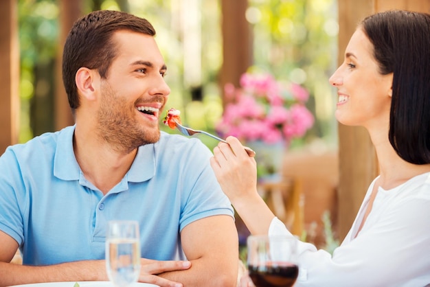 Try my meal! Beautiful young woman feeding her boyfriend with salad and smiling while both relaxing in outdoors restaurant