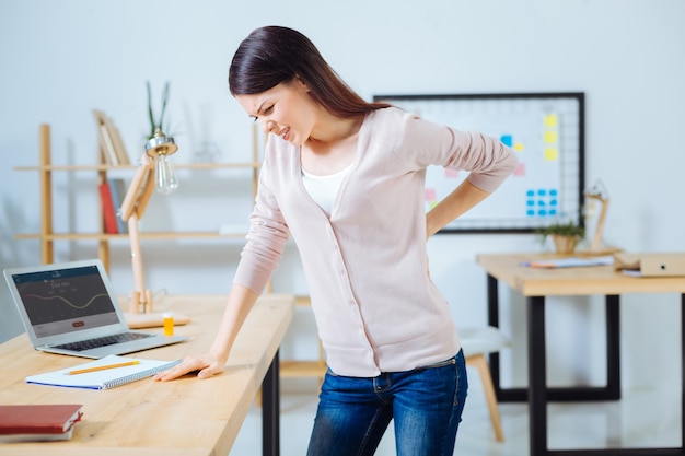 Try to undergo. Sick woman standing near her workplace and leaning on table while looking downwards