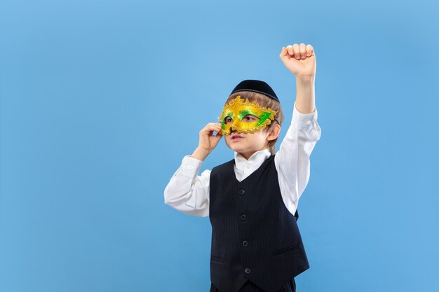 Trying carnival mask on. Portrait of a young orthodox jewish boy isolated on blue studio wall.