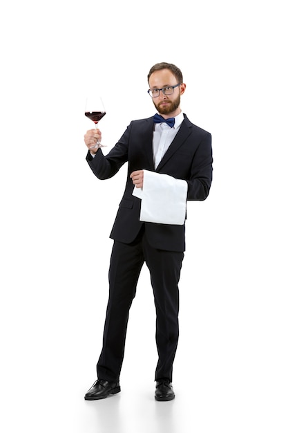 Trying red wine on. Portrait of male sommelier, wine steward or bar worker in white and black suit isolated over white background. Copyspace for ad. Concept of professional occupation, job.