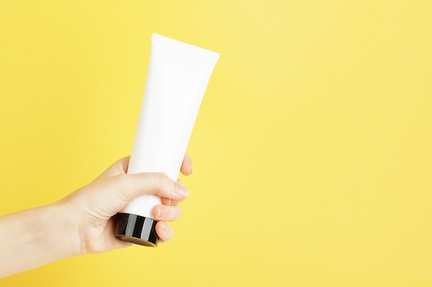 A tube of cream in a white package with a black cap holds in female hand on a yellow background. Lotion, body cream, skin care in a plastic container. Mock up, copy space. Summer skincare concept.