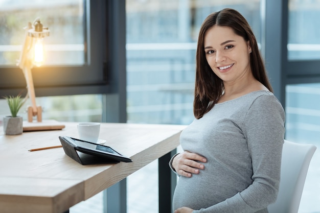 Tue beauty. Close up of gracious pregnant woman looking at you with a smile while sitting at the table and touching her stomach