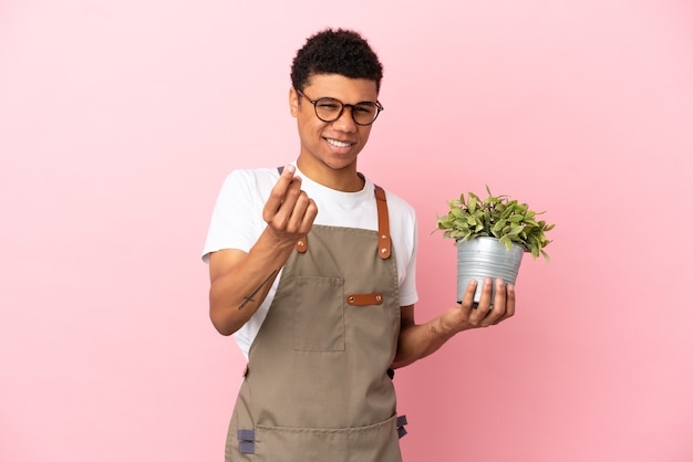 Tuinman Afrikaanse man met een plant geïsoleerd op roze achtergrond geld gebaar maken