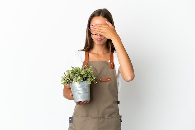 Tuinman meisje met een plant over geïsoleerde witte achtergrond die ogen bedekt door handen. Wil je iets niet zien