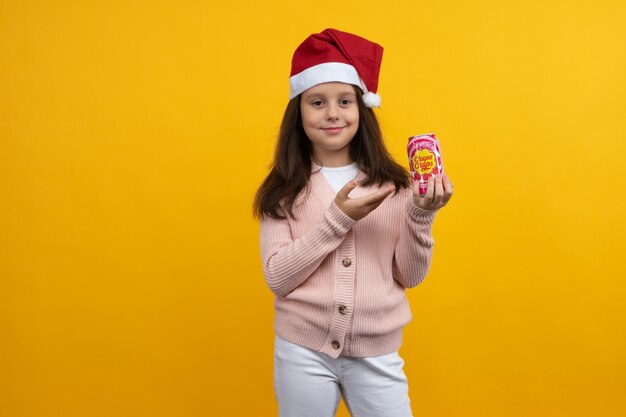 Tula, rusland - 23 augustus 2021: een jong meisje in een kerstmuts van de kerstman met een pot chupa chups drinkt in haar handen op een gele achtergrond