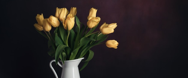 Tulpen in een kruik. Klassiek stilleven met een boeket gele tulp bloemen in een vintage witte kruik op een donkere muur en een oude houten tafel.