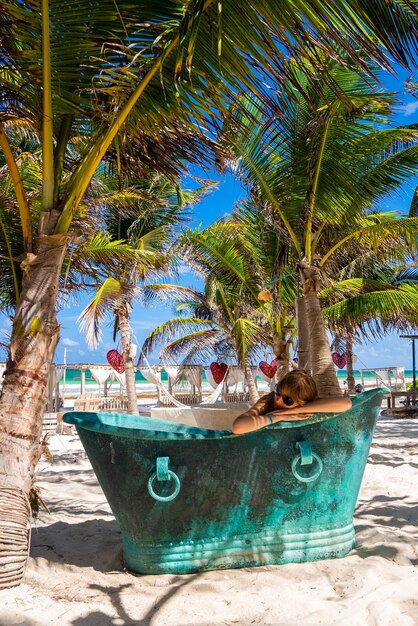 Tulum, Mexico. May 10, 2021. Woman relaxing in retro vintage style metallic bath tub on sand at beautiful beach resort in tulum, Mexico.