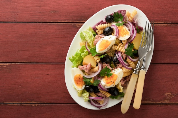 Foto insalata di tonno con pasta uova patate olive cipolle rosse e salsa in piatto bianco su sfondo vecchio tavolo rustico rosso insalata nizzarda cucina francese vista dall'alto posa piatta