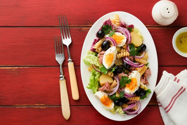 Tuna salad with pasta eggs potatoes olives red onions and sauce in white plate on old red rustic table background nicoise salad french cuisine top view flat lay