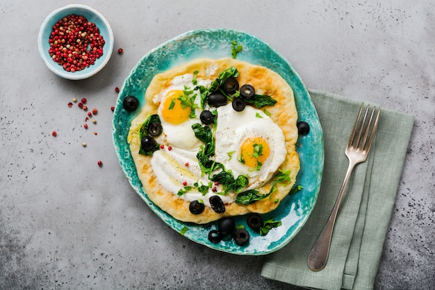 Turkish eggs flatbread with yoghurt, cheese, olives, spinach and red pepper on ceramic vintage plate on gray old surface. Top view.