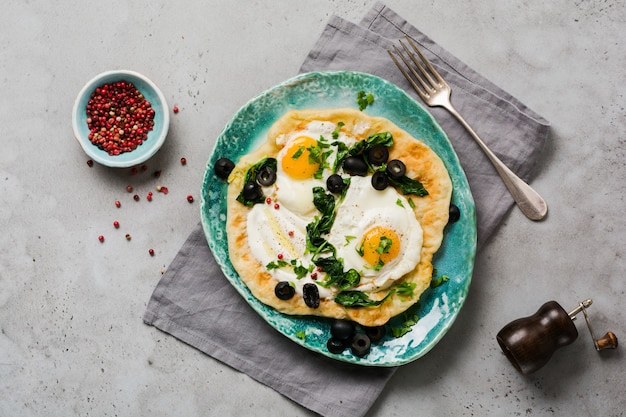 Turkish eggs flatbread with yoghurt, cheese, olives, spinach and red pepper on ceramic vintage plate on gray old surface. Top view.